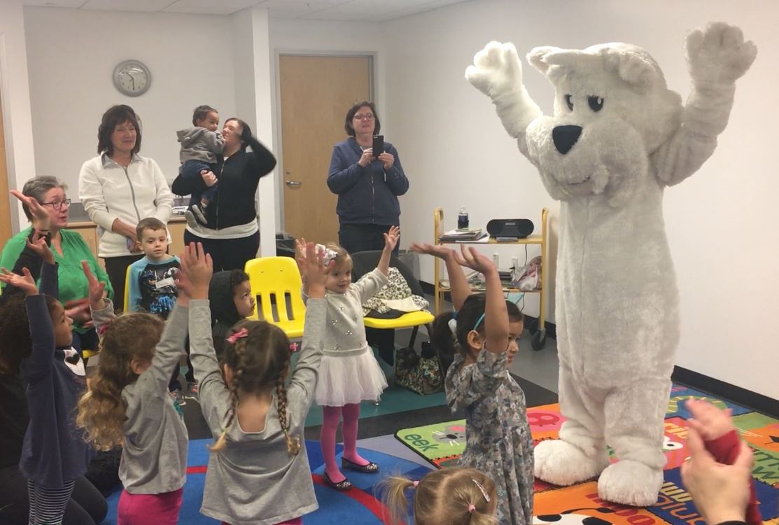 Image of the city mascot, a large white dog named Romi, with kids at the library.