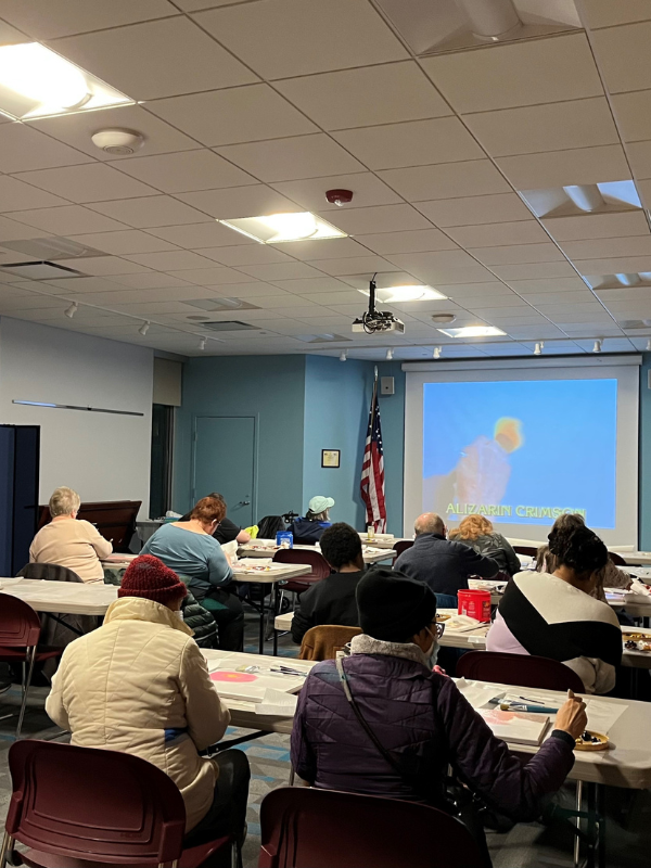 A group of people paint along to a video tutorial on a screen.
