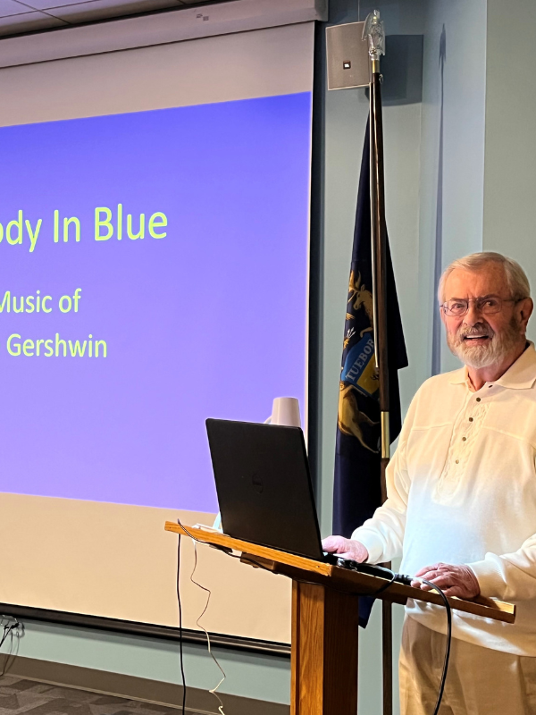 Presenter stands beside a screen that reads: Rhapsody in Blue: The Music of George Gershwin.
